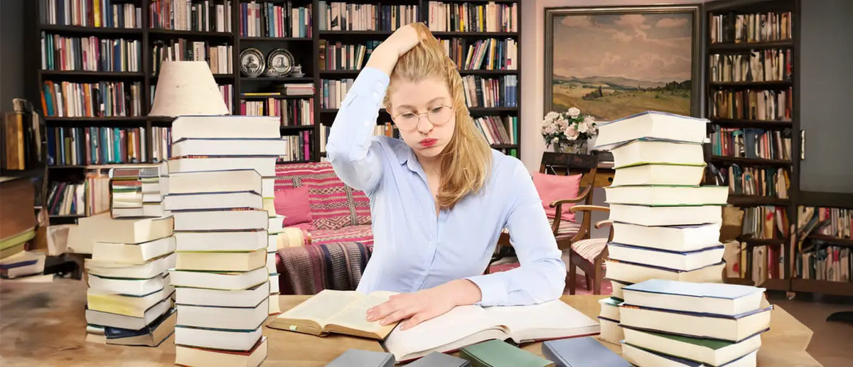 Eine junge Frau die zwischen Stapeln von Büchern sitzt und sich mit der Hand angestrengt durch die Haare fährt.