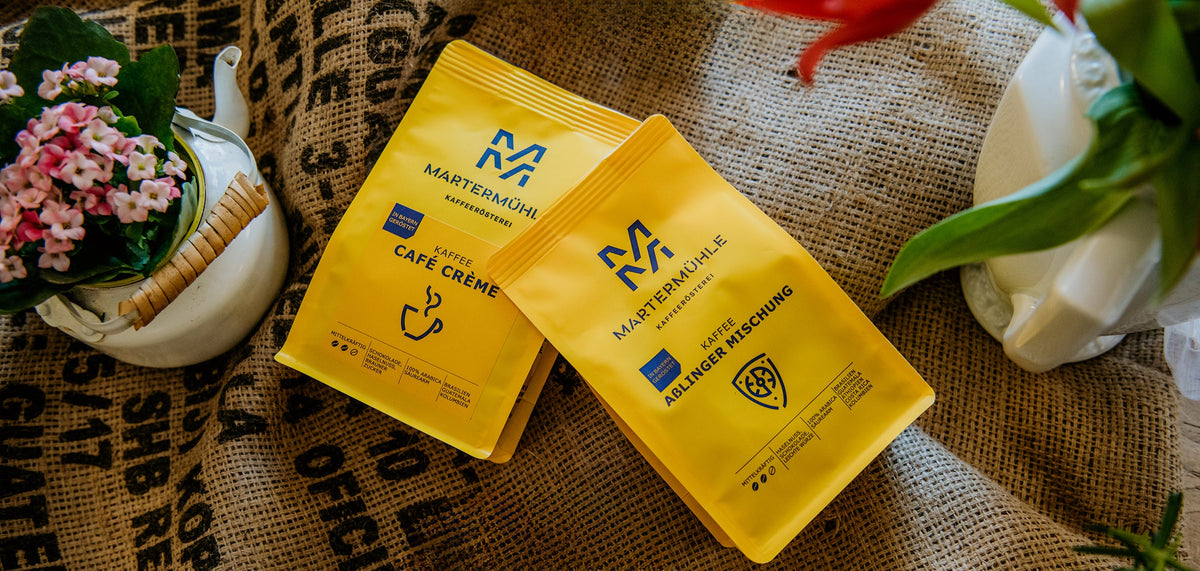 Zwei gelbe Tüten Kaffee der Kaffeerösterei Martermühle und Blumen liegen auf Kaffeesäcken